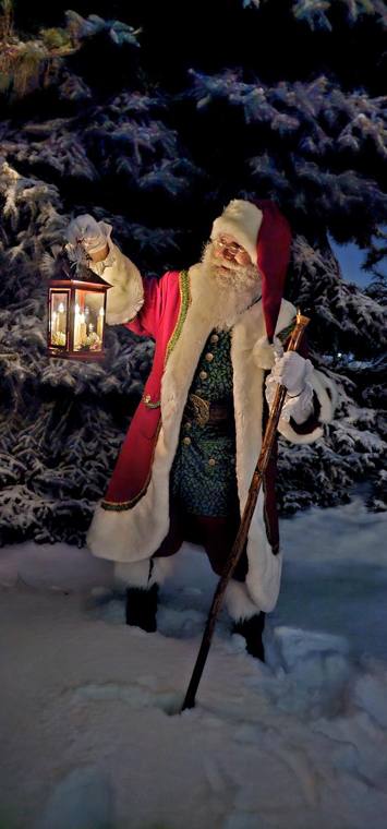 Santa Phil with a lit lantern and walking staff among snow-covered pines at night